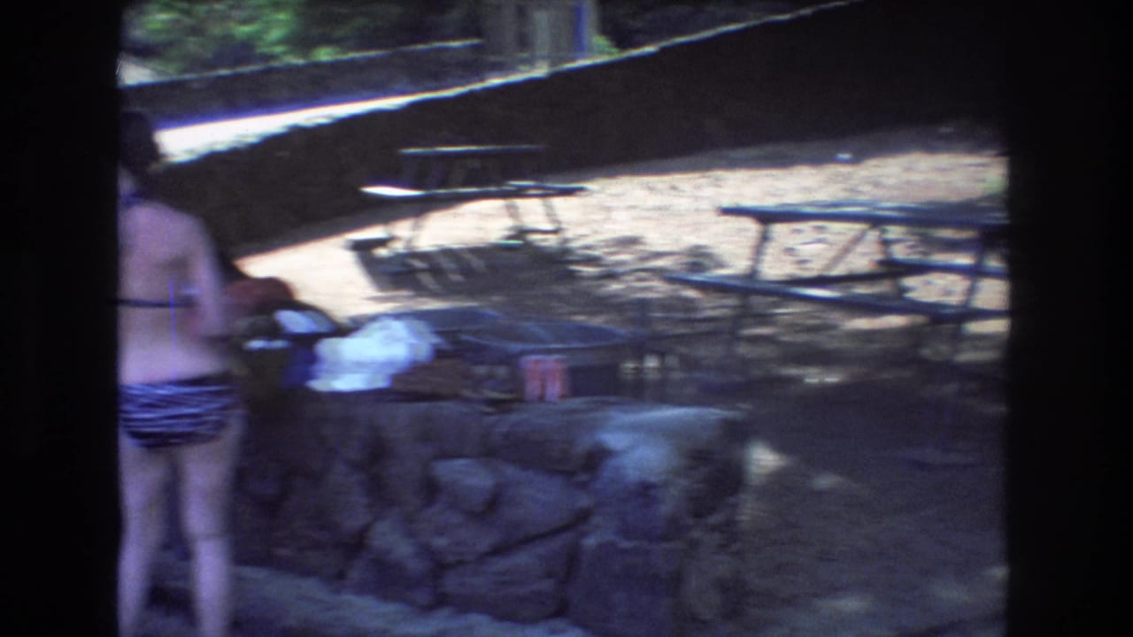 In a 1977 Honolulu home-movie frame, a woman in a striped bikini and a partially visible man stand near stone picnic structures, grills, tables, and trees.