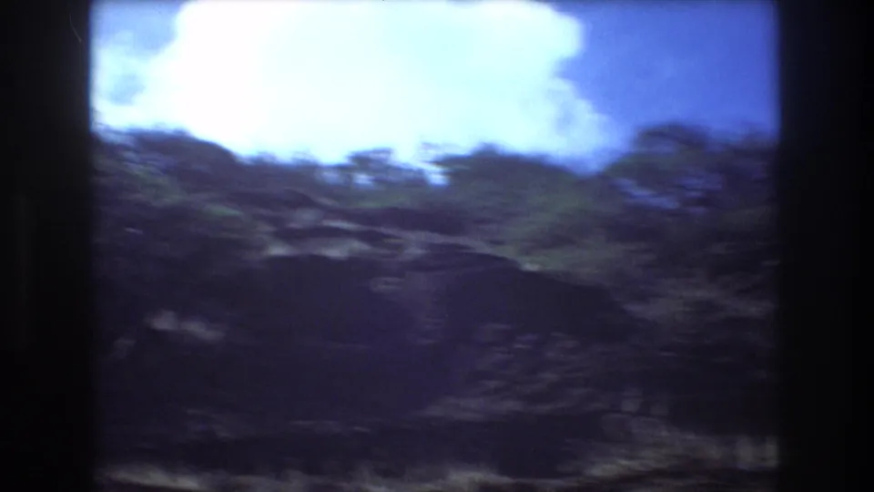 Handheld 8mm view in Honolulu showing a motion-blurred vegetated slope below a bright white cloud and blue sky.