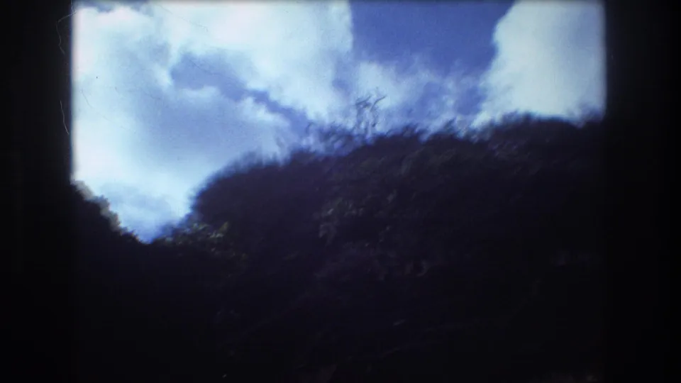 1977 Honolulu home-movie frame with dark hillside and brush rising into a bright cloudy sky, softened by exposure and motion.