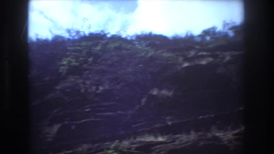 Handheld 8mm frame of a steep Honolulu hillside with dark rocky areas, green vegetation, and bright sky along the top edge.