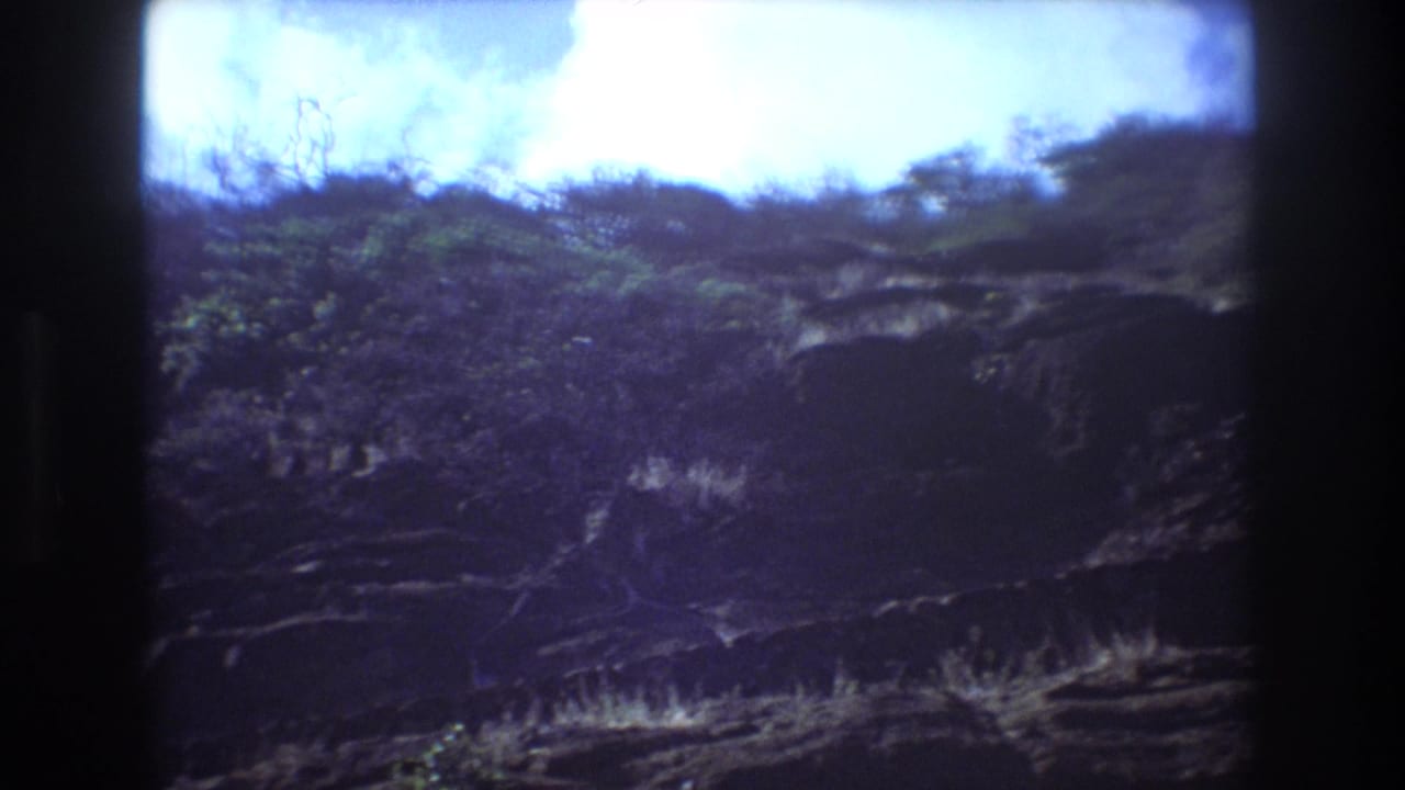 1977 Honolulu home-movie frame showing a rugged rocky hillside with scrub vegetation beneath a bright sky, bordered by dark 8mm film edges.