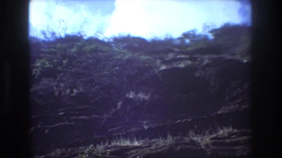 1977 Honolulu home-movie frame showing a rugged rocky hillside with scrub vegetation beneath a bright sky, bordered by dark 8mm film edges.