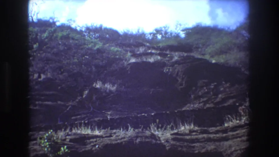 1977 Honolulu 8mm frame with a rocky slope, scattered green growth, and bright overexposed sky across the top of the frame.
