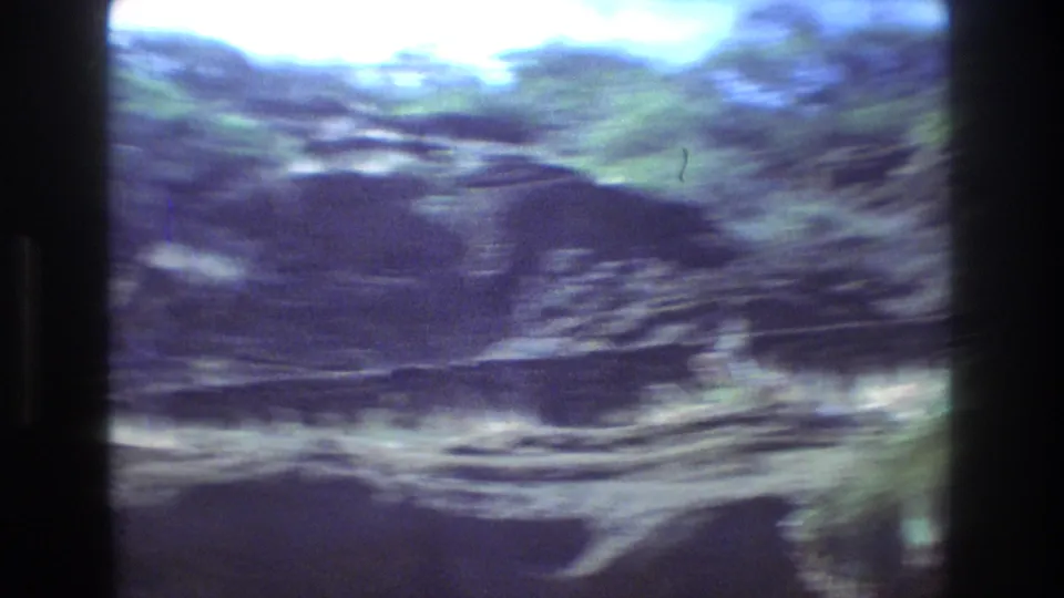 Motion-blurred 1977 Honolulu home-movie frame showing a pan across rocky terrain, scrub vegetation, and a bright sky.