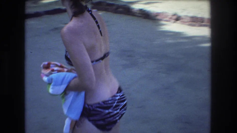 1977 Honolulu home-movie frame showing a woman in a striped swimsuit carrying towels while walking through a sunlit tropical outdoor area.