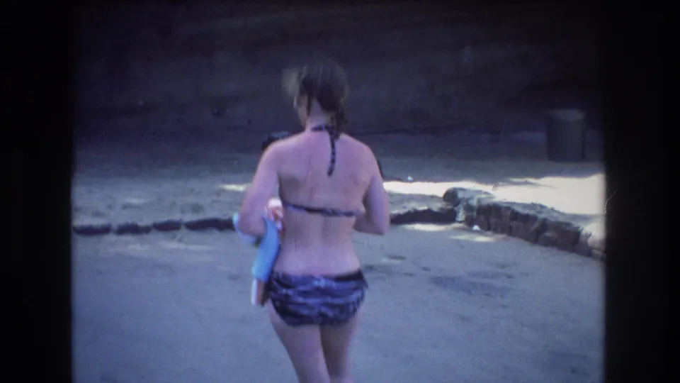 1977 Honolulu 8mm frame showing a woman seen from behind in a striped swimsuit, carrying towels while walking across a sandy area bordered by low stones.