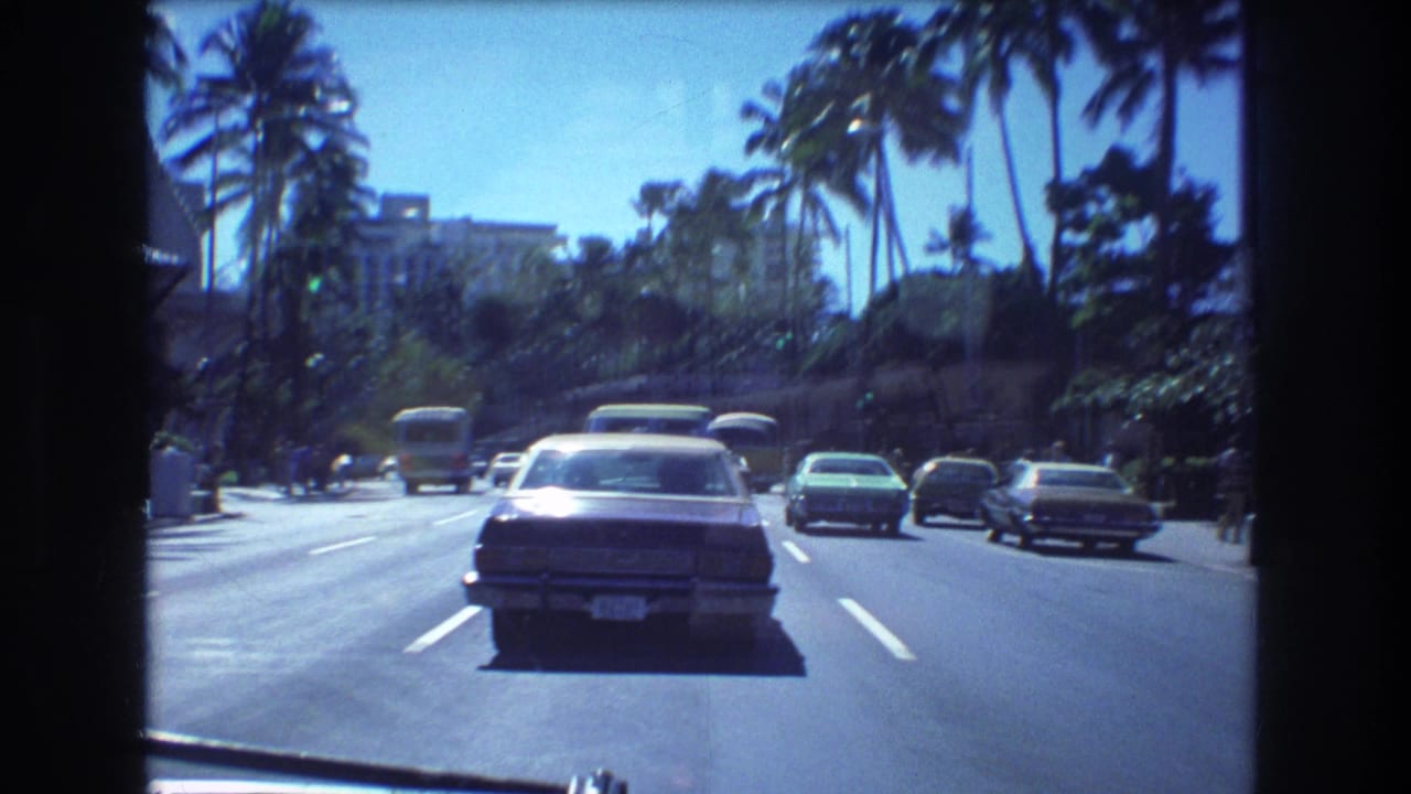 1977 Honolulu 8mm frame showing a passenger-view drive along a sunny palm-lined boulevard with a sedan ahead, other cars, a bus, and distant buildings.