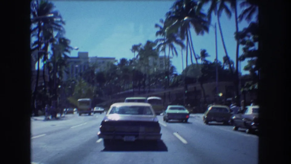 1977 Honolulu street scene from inside a moving car, with tall palms, a sedan in the center lane, a green car, buses, and distant urban buildings.