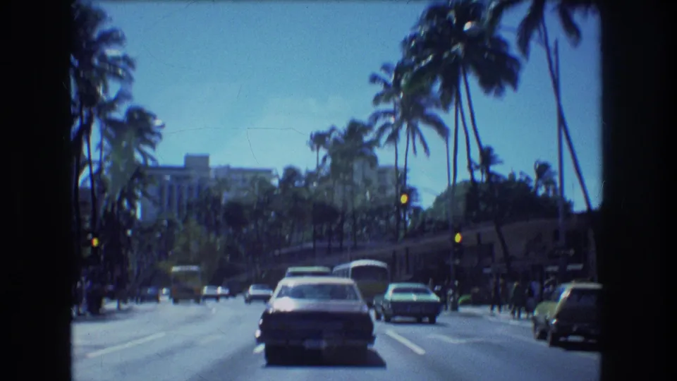 1977 Honolulu 8mm frame with a windshield-level view of traffic proceeding down a palm-lined boulevard, including cars, traffic lights, sidewalks, and background buildings.