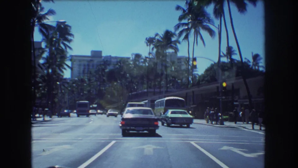 1977 Honolulu street footage showing cars and buses near an intersection on a palm-lined boulevard, with small distant pedestrians along the roadside.