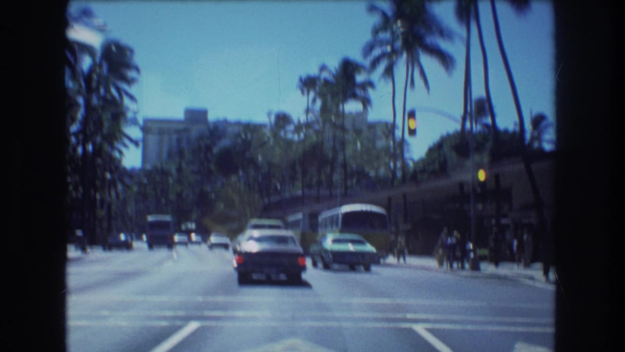 Cars drive along a palm-lined city street with a bus and distant building visible.