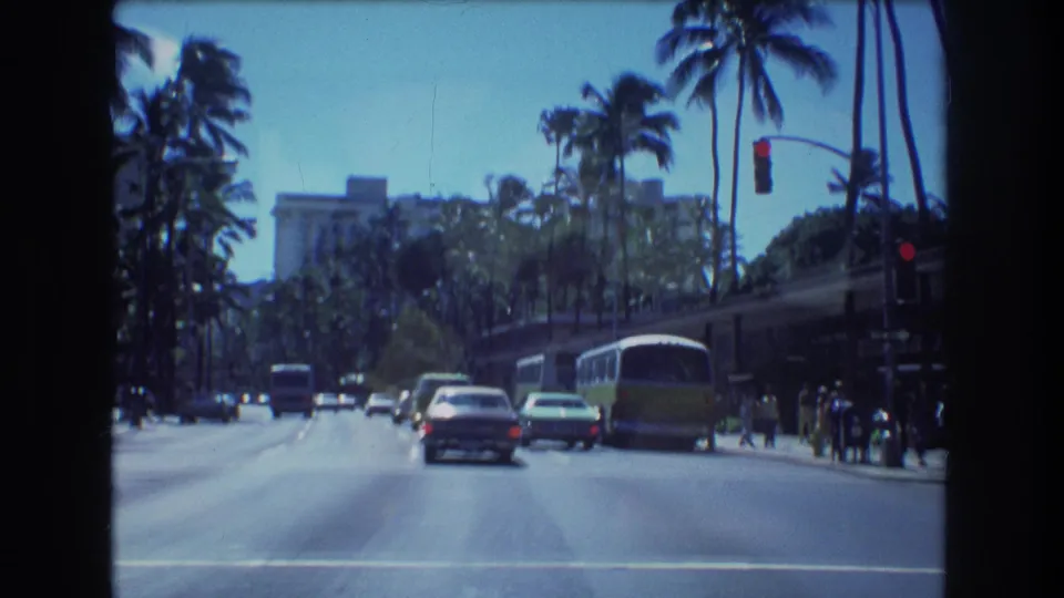 1977 Honolulu street view with a green-and-white bus on the right, cars ahead, red traffic signal, pedestrians, palms, and a large building in the background.