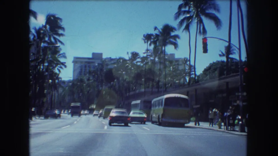 1977 Honolulu 8mm frame showing moving cars, a bus near the right side of the road, pedestrians at the sidewalk, tall palms, traffic signals, and a distant resort-like building.
