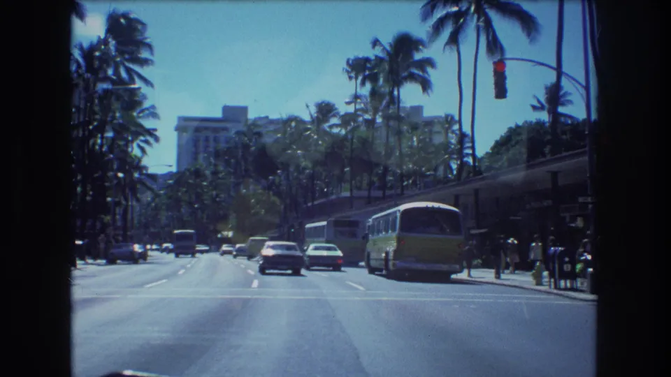 1977 Honolulu driving shot with older cars, a green-and-white bus, red traffic lights, tall palms, distant pedestrians, and a large building beyond the trees.