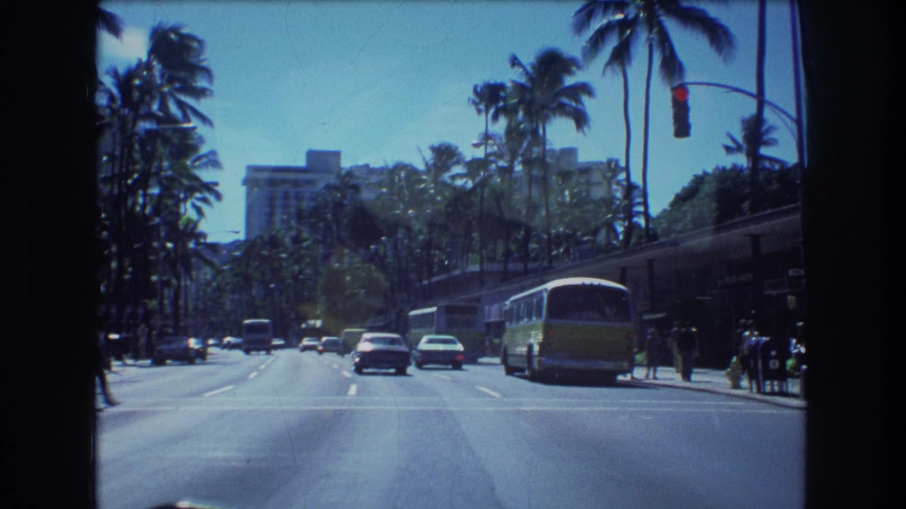 1977 Honolulu street view from a moving vehicle, with palm trees, cars, a yellow-and-white bus, traffic lights, and pedestrians near a marked crosswalk.