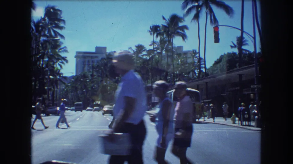 1977 Honolulu home-movie frame with blurred pedestrians in the foreground and cars, palm trees, and a city street behind them.