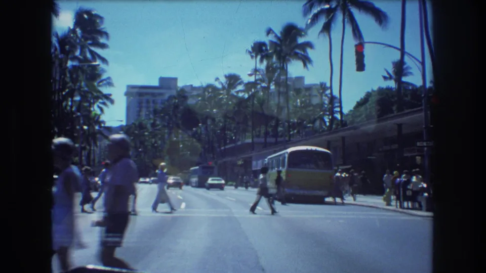 1977 Honolulu street scene showing pedestrians in a marked crosswalk, a yellow-and-white bus, cars, palm trees, and bright midday light.