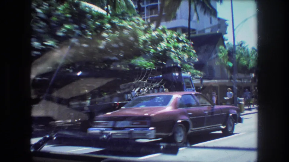 1977 Honolulu home-movie frame looking through a car windshield at a maroon sedan, a blue tow truck, pedestrians, trees, and urban building facades.