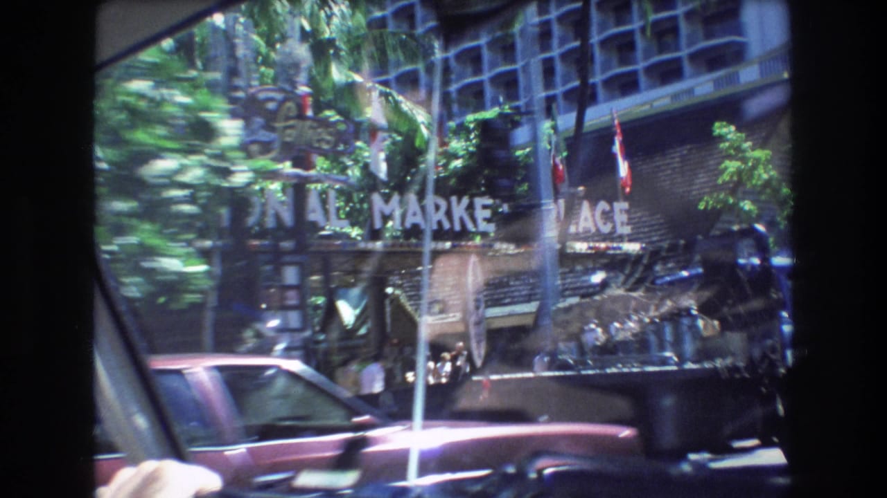1977 Honolulu street view from inside a moving car, with the Market Place sign, flags, tropical trees, and a maroon vehicle visible through windshield reflections.