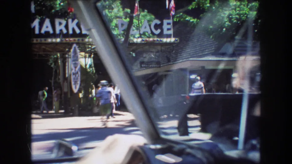 1977 Honolulu home-movie frame showing pedestrians near a Market Place entrance, filmed through a car window with strong windshield framing and street reflections.
