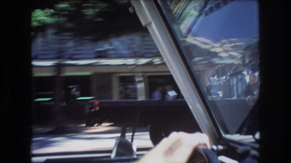 1977 Honolulu driving view from inside a car, with a shaded building, angled windshield frame, blurred traffic, and bright reflections from the moving street scene.