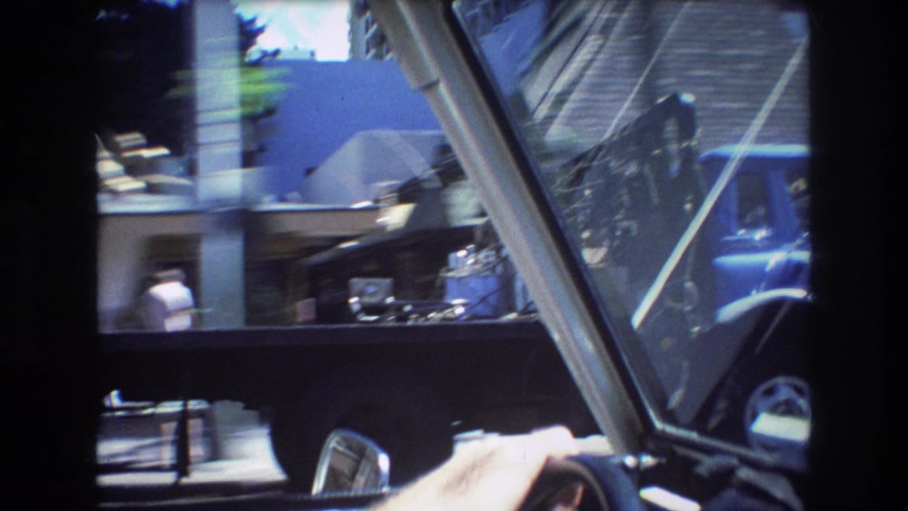1977 Honolulu home-movie driving POV through a windshield, showing the steering wheel, a hand, a blue tow truck, and passing roadside structures.