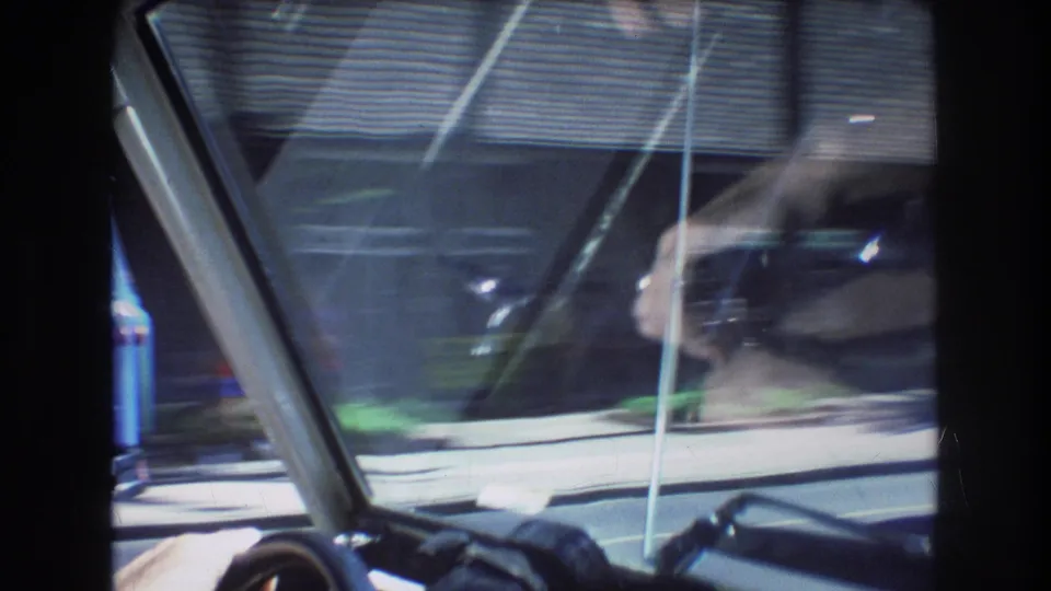Soft 1977 Honolulu driving POV through the windshield, with steering wheel detail, passing street, trees, and blurred building forms.