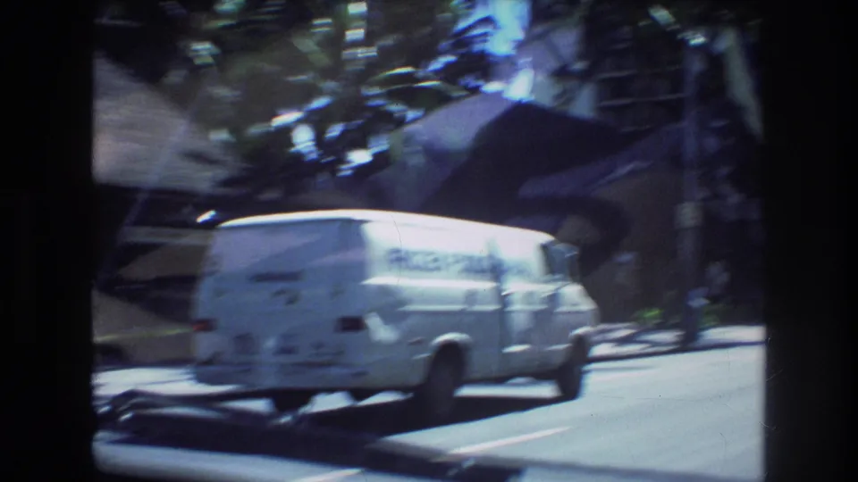 A white delivery van moves through a sunlit Honolulu street scene from 1977, with palm trees, roadside greenery, and high-rise buildings in soft 8mm focus.