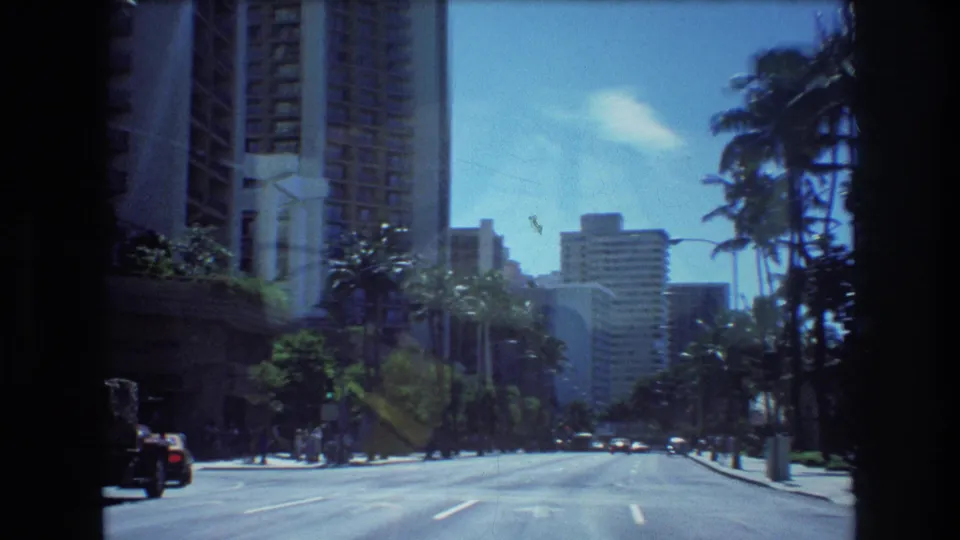 The camera looks ahead along a Honolulu street in 1977, showing palms, high-rise buildings, distant cars, sidewalks, and pedestrians under a bright blue sky.