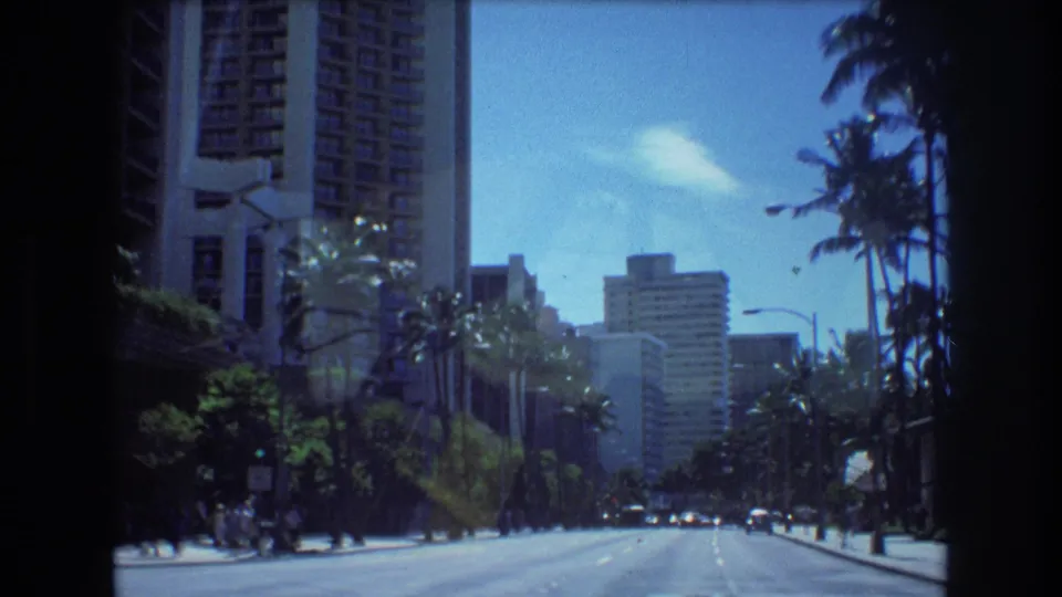 A wide 1977 Honolulu roadway extends toward distant buildings, with palm trees, high-rises, small vehicles, and soft 8mm grain visible across the frame.