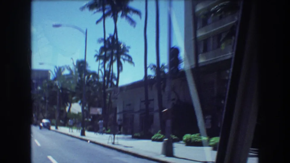 1977 Honolulu street footage showing a road, sidewalk, tall palms, streetlights, distant vehicles, and a large building edge filmed from a moving car.