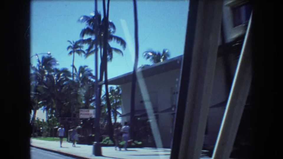 1977 Honolulu home-movie frame with pedestrians on a sidewalk, tall palm trees, a roadside sign, and a large building passing close to the vehicle.