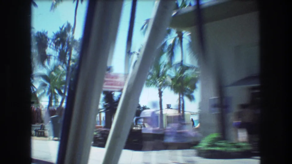 1977 8mm car-window view in Honolulu with motion-blurred palms, sidewalk, building columns, parked or passing vehicles, and bright midday light.
