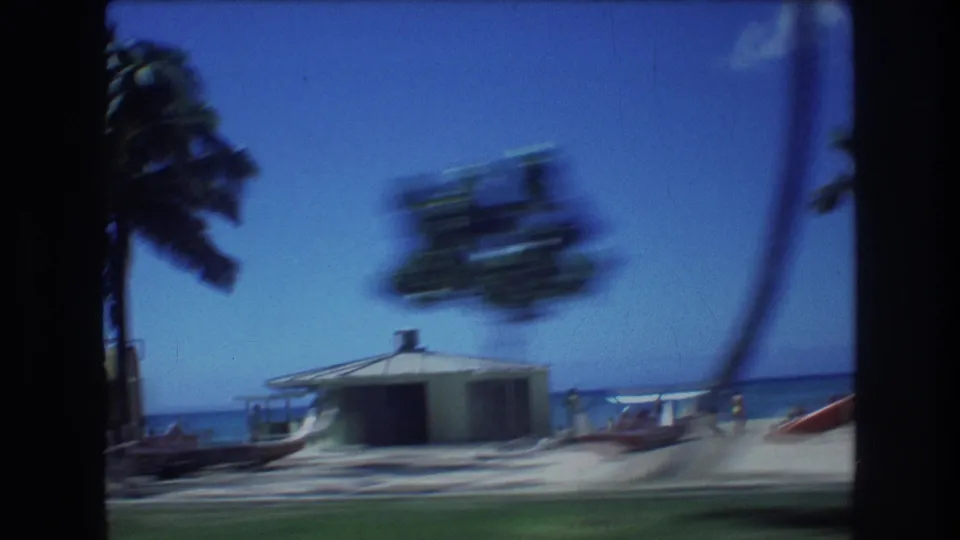 1977 Honolulu drive-by frame with a small shore building, palm trees, ocean, and surfboards or beach craft visible beyond the roadside foreground.