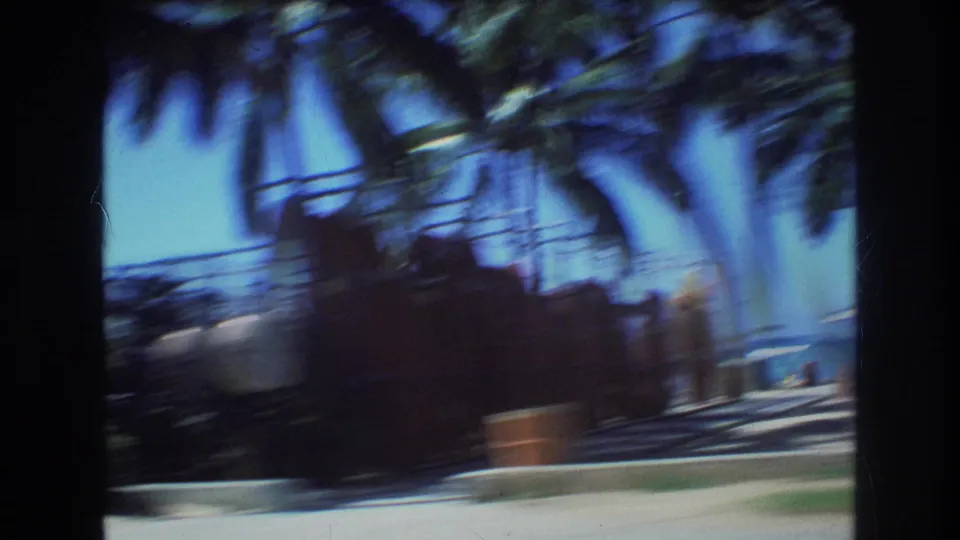 1977 Honolulu home-movie frame with heavy motion blur across palms, beach equipment, shoreline, and ocean as the camera passes by from inside a car.