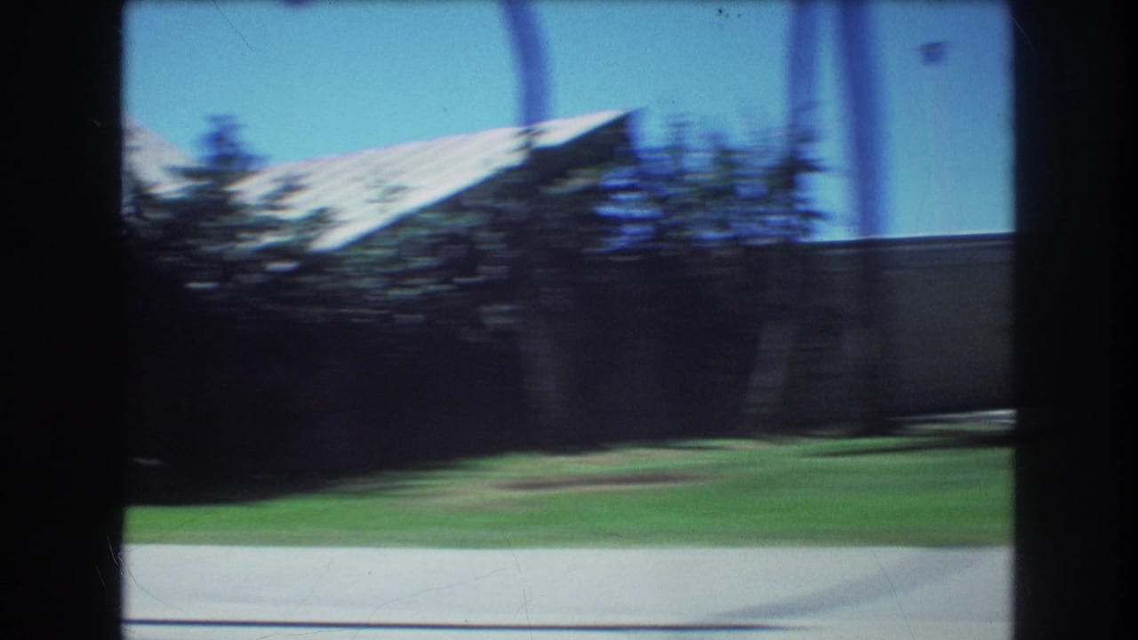 1977 Honolulu 8mm car-window view with dark interior dividers framing blurred roadside grass, trees, a low wall, and a pale angular roofline under bright blue sky.
