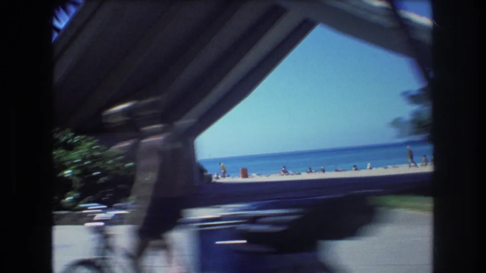 1977 Honolulu beach and blue ocean seen through an angular archway or canopy from a moving car, with small beachgoers in the distance and a dark vehicle blurred in front.
