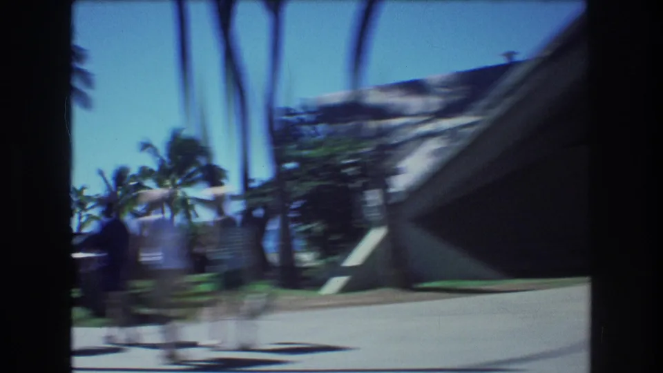 1977 Honolulu moving-car frame with blurred pedestrians, palm trees, roadside grass, and an angular beachside structure visible through dark car-window framing.