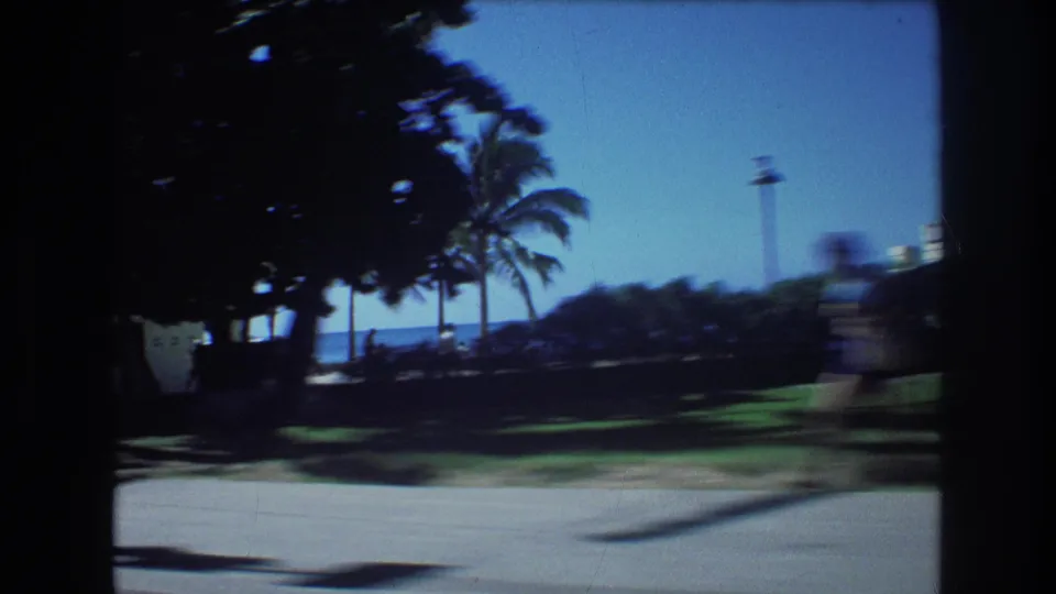1977 Honolulu 8mm frame showing palms, shaded beachside lawn, a strip of blue ocean, and a blurred pedestrian near the right edge from a moving car perspective.