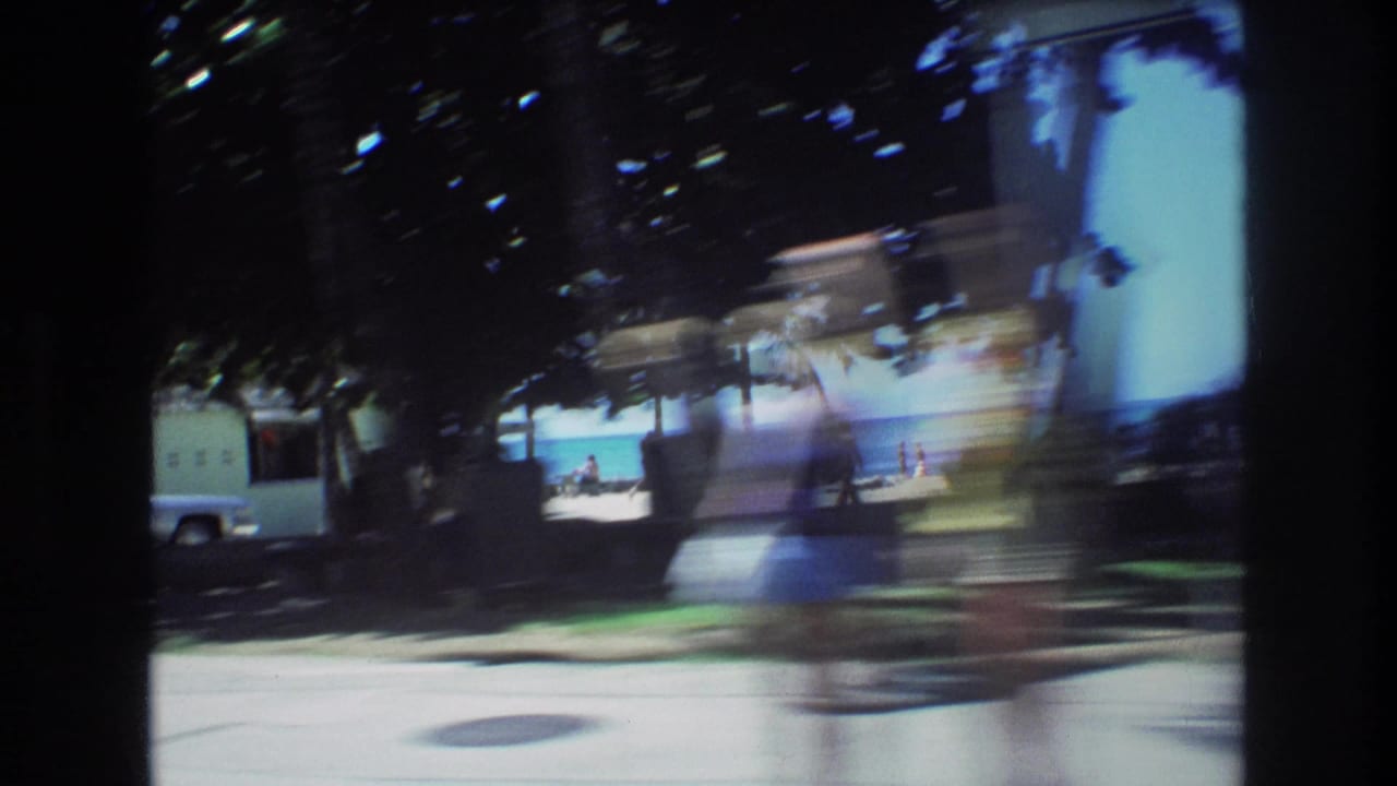 1977 Honolulu home-movie frame from a moving vehicle, showing shadowed trees, bright pavement, beach frontage, and distant figures near the ocean.