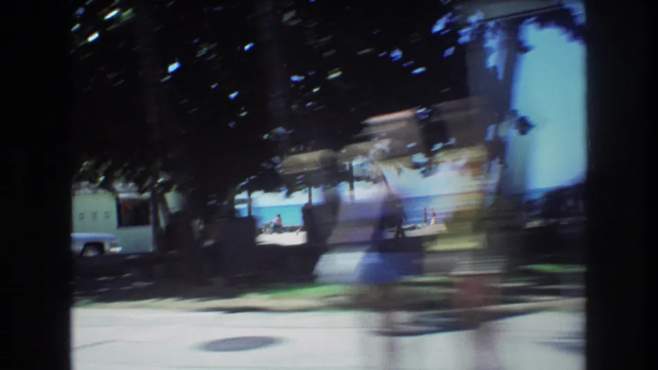 1977 Honolulu home-movie frame from a moving vehicle, showing shadowed trees, bright pavement, beach frontage, and distant figures near the ocean.