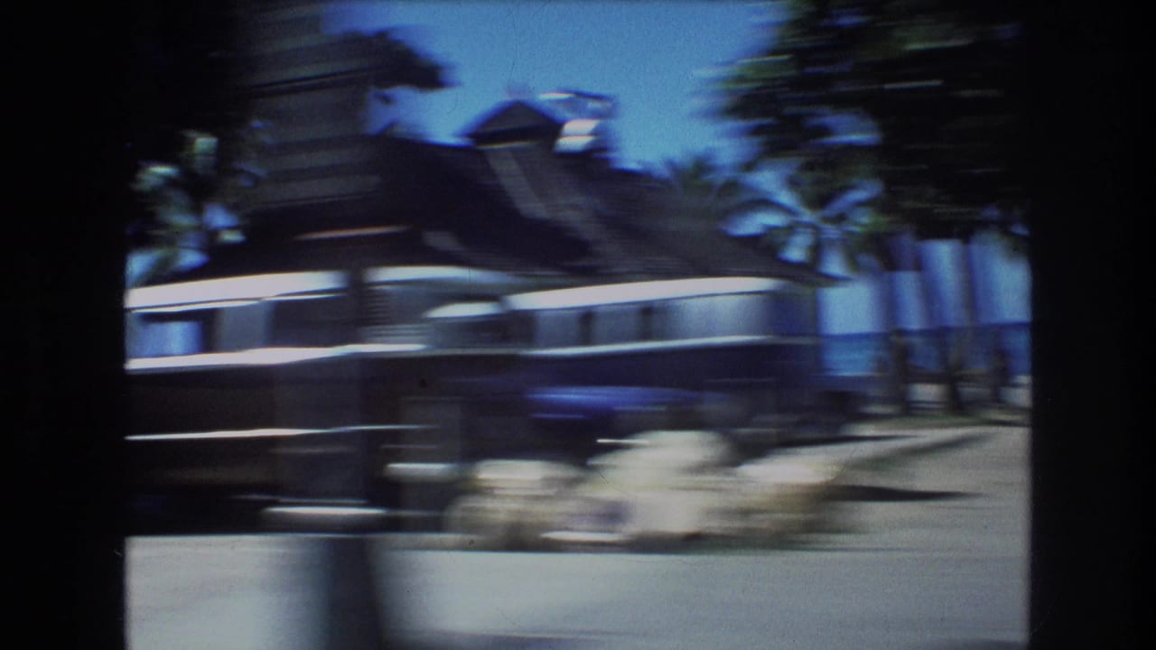 Motion-blurred roadside view of a dark building, palm trees, sidewalk, and bright blue sky.