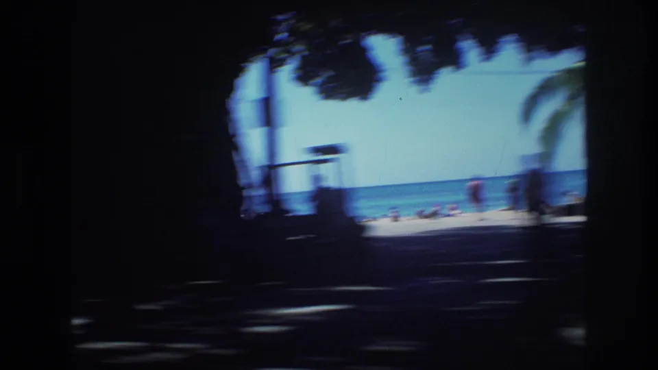 1977 Honolulu home-movie frame with dark tree shadows in the foreground, a bright beach strip, blue ocean, and distant beachgoers.