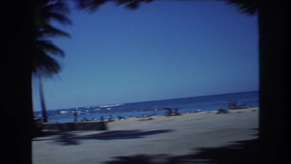 1977 Honolulu beach frame showing a palm tree at left, open sand, ocean horizon, and small distant groups of beachgoers under a clear blue sky.