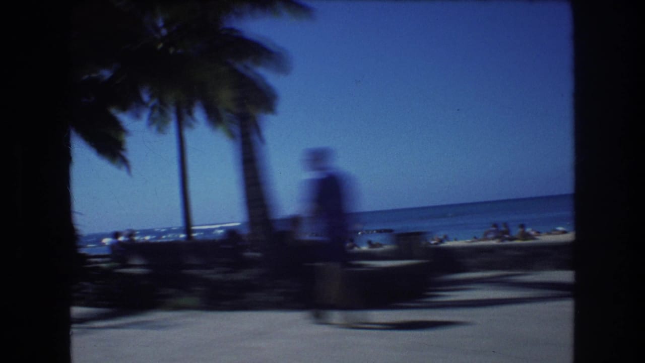 1977 Honolulu home-movie frame showing a palm-lined paved beachfront walkway, blue ocean horizon, beach sand, and blurred people moving through the scene.
