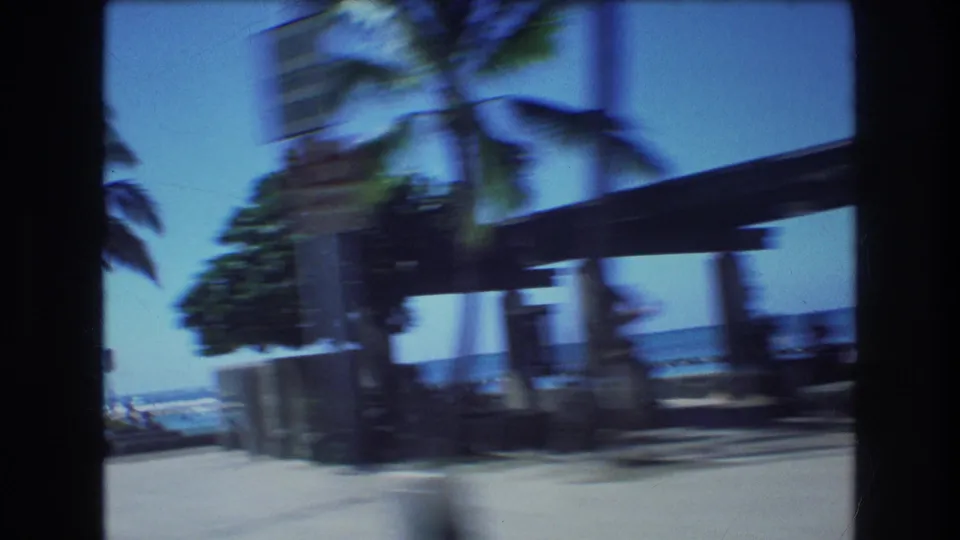 Handheld 1977 Honolulu beach frame with motion-blurred palm trees, a covered structure, ocean beyond the walkway, and faint distant figures near the shore.