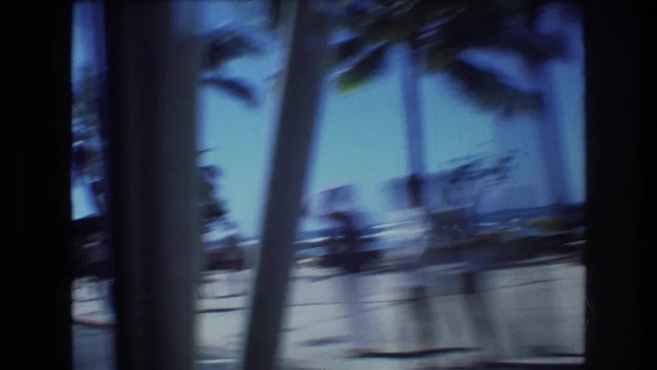 1977 Honolulu 8mm frame with blurred palm trunks in the foreground, paved seaside walkway, beach, ocean horizon, and indistinct people near the shore.