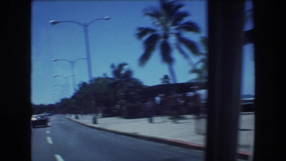 1977 Honolulu road view from a moving vehicle, showing a palm-lined multi-lane street, classic cars, streetlights, and hotel buildings under bright sky.