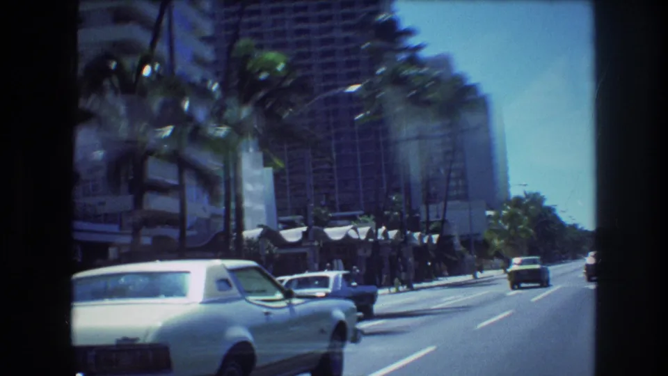 1977 Honolulu street scene with a white classic car, palm trees, pedestrians near hotel frontage, and tall urban resort buildings visible from a moving vehicle.