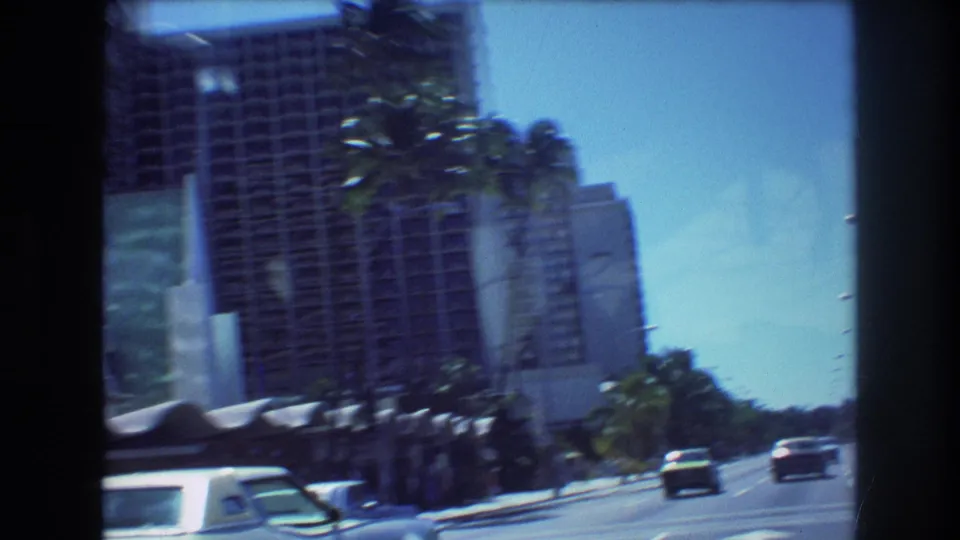 Motion-blurred 1977 Honolulu street view with hotel towers, palm trees, classic cars, and pedestrians along the roadside.